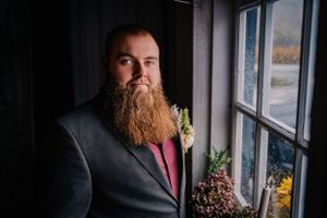 Close-up side profile of person with long beard wearing dark suit jacket by window.