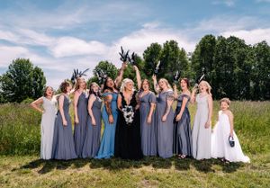 Bridesmaids in coordinated dresses make silly gestures for a group photo in a meadow.