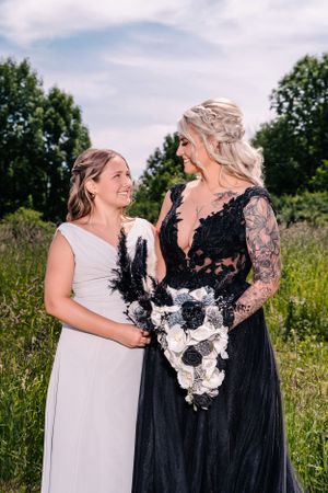 Two people in formal dresses share a touching moment in a sunlit meadow while holding black and white floral bouquets.