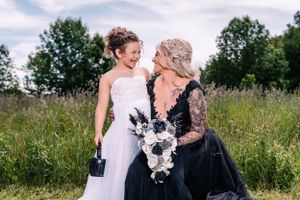 Two people share laughter while wearing formal dresses and holding bouquets in a summer meadow.