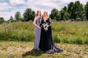 Two people in elegant dresses stand together in a meadow holding black and white floral arrangements.