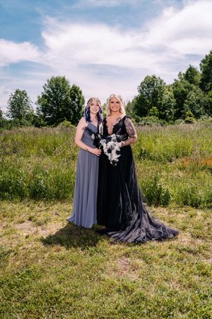 Two figures in elegant gowns pose together in a lush green field under blue skies.