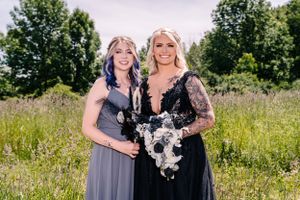 Two women in formal attire stand together in a wildflower field holding bouquets.