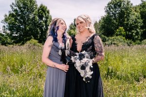 Two people in elegant dresses laugh together while holding bouquets in a meadow.