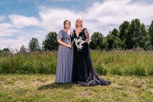 Two figures in elegant dresses pose with bouquets against a cloudy summer sky.