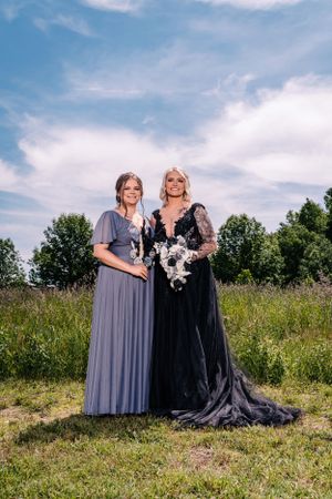 Two ladies stand together in a field wearing floor-length gowns against a blue summer sky.