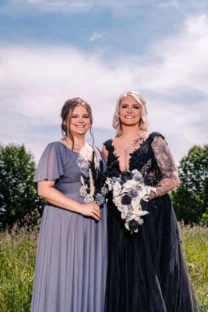 Two women pose in elegant formal gowns, one in gray and one in black with lace details, standing in a sunny meadow.