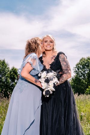 Two people embrace while wearing formal dresses in a sunny field with trees in the background.