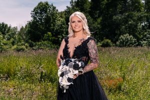 Person in black lace gown holds white floral bouquet while standing in sunny meadow.