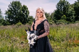 Person poses with white bouquet wearing black lace gown in sunny outdoor setting.