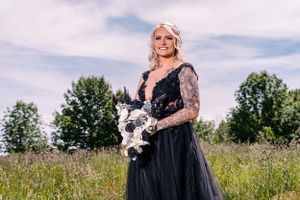 Person in black lace wedding gown holds white bouquet while standing in grassy field.