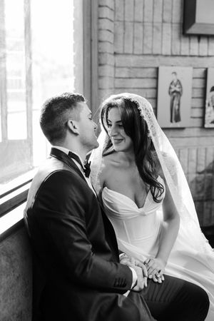 Black and white portrait of smiling bride in veil and groom in tuxedo sharing tender moment by window.