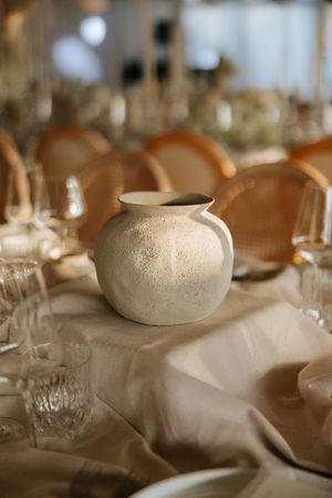 Rustic white ceramic vase centerpiece on elegant beige linen wedding reception table with crystal glasses.