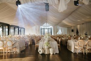 Elegant all-white wedding reception tent with draped ceiling, long tables, gold chairs, and soft lighting.