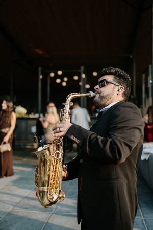Saxophonist in sunglasses and dark suit performs at outdoor wedding cocktail hour with string lights behind.