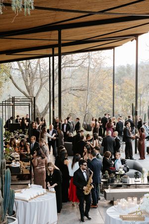 Elegant wedding cocktail hour reception with guests mingling under a modern open-air pavilion surrounded by autumn trees.