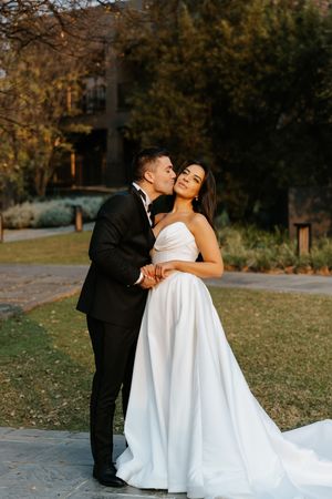Groom in black tuxedo kisses bride in strapless white ball gown outdoors at golden hour.