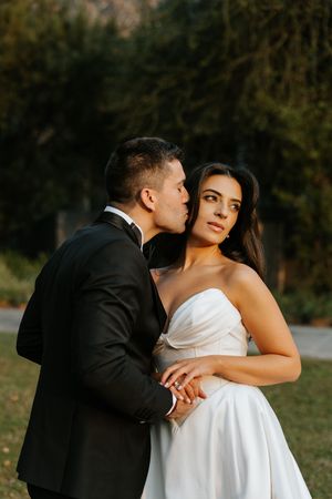 Groom in black tuxedo kisses bride in strapless white gown during romantic outdoor wedding portrait.