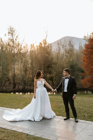 Bride and groom hold hands on stone path with golden autumn trees and mountains at sunset.