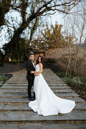 Bride and groom pose on stone steps surrounded by trees in elegant black tuxedo and white gown.
