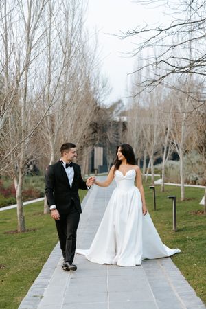 Bride and groom walk hand in hand along garden pathway lined with bare trees at wedding venue.