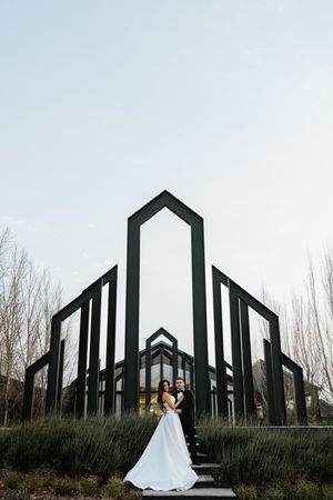 Bride and groom embrace before dramatic black geometric arch structure at modern outdoor wedding venue.
