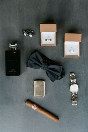 Groom wedding flat lay featuring cologne, cufflinks, bow tie, watch, lighter, cigar, and two ring boxes.