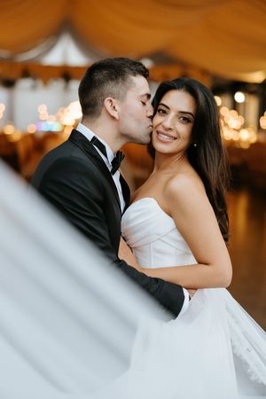 Groom kisses smiling bride on cheek inside warmly lit wedding reception tent with bokeh lights.