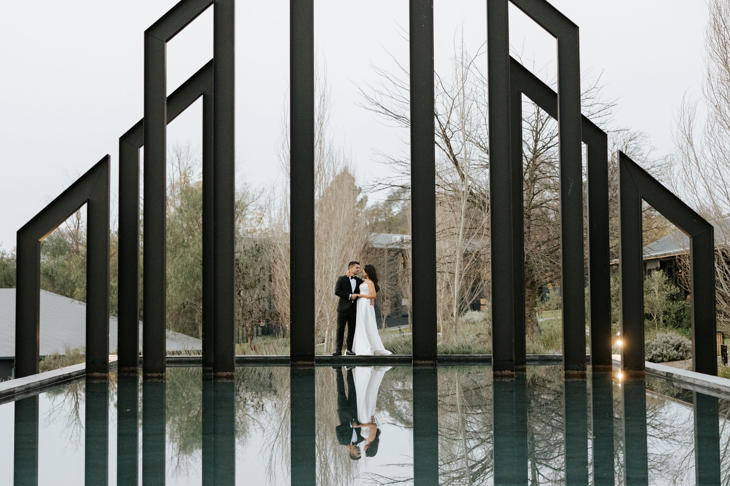 Bride and groom embrace between modern black geometric frames reflected in still water at wedding venue.