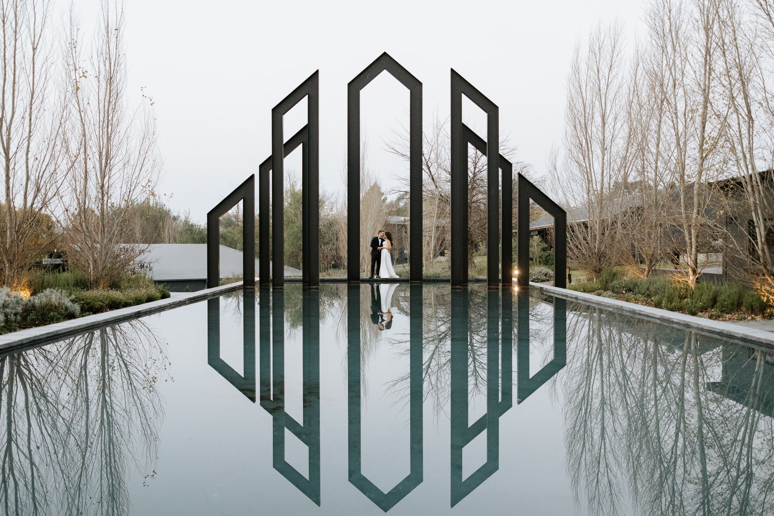 Couple embracing between geometric black frames reflected in still water at an elegant outdoor wedding venue.