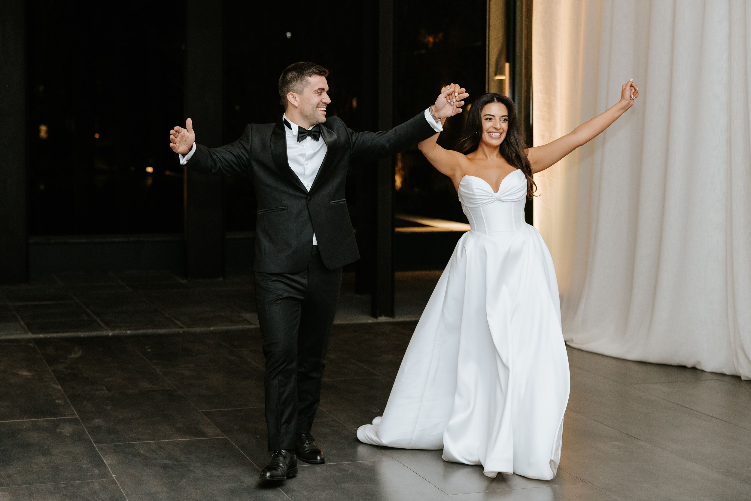 Bride and groom make grand entrance at wedding reception, raising arms in celebration wearing white gown and black tuxedo.