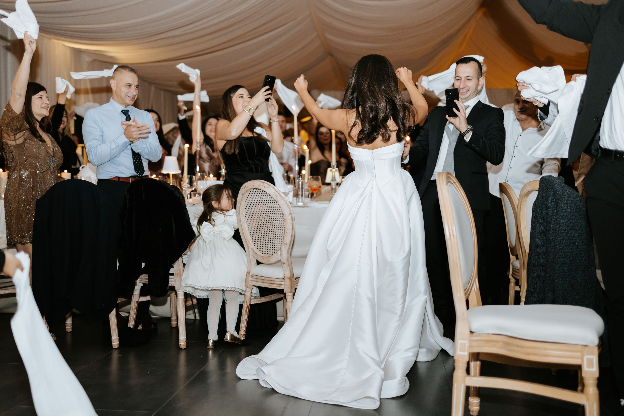 Wedding guests wave white napkins in celebration around bride in ballgown at elegant tented reception venue.