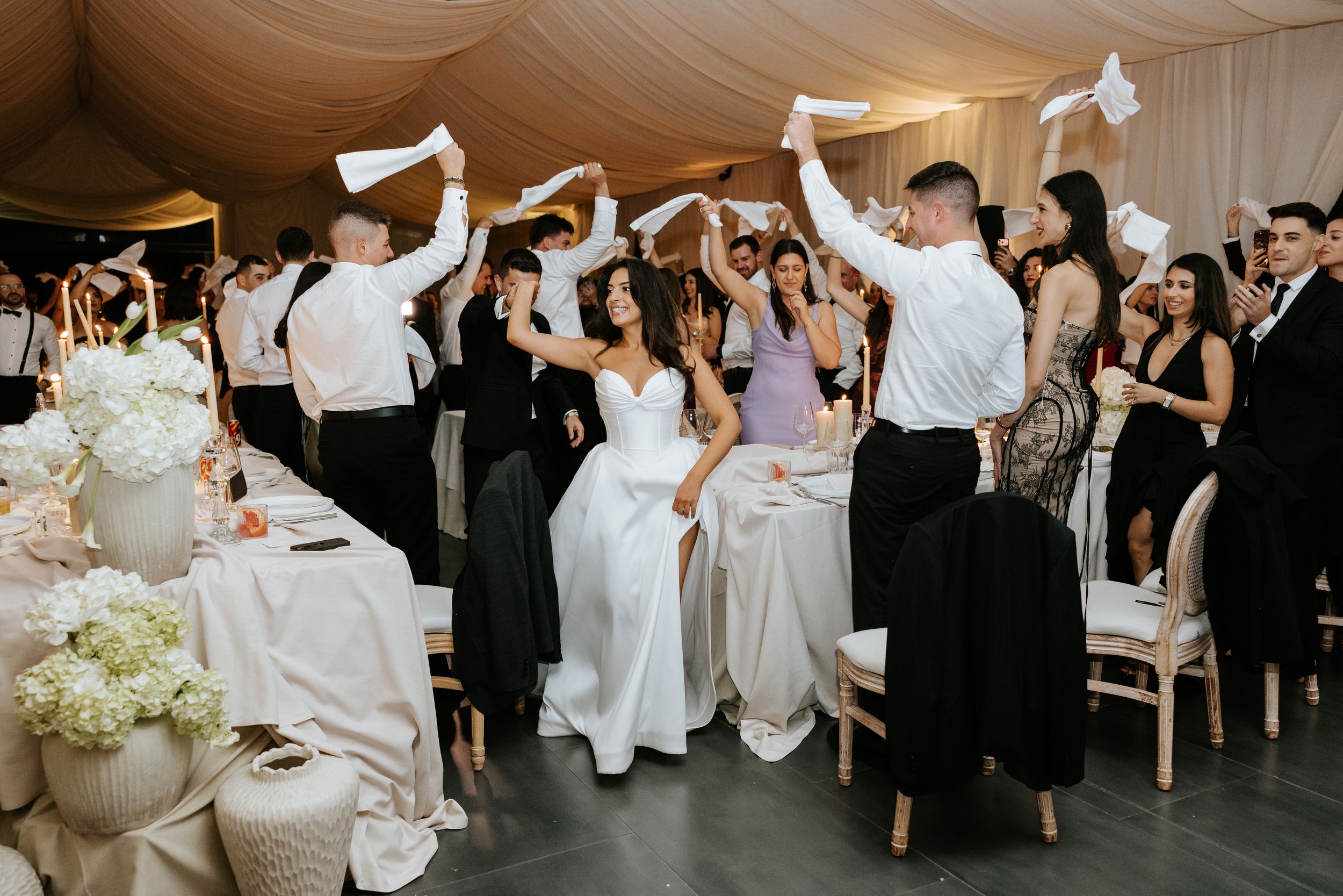 Joyful wedding guests wave white napkins surrounding bride in strapless gown during lively reception celebration.