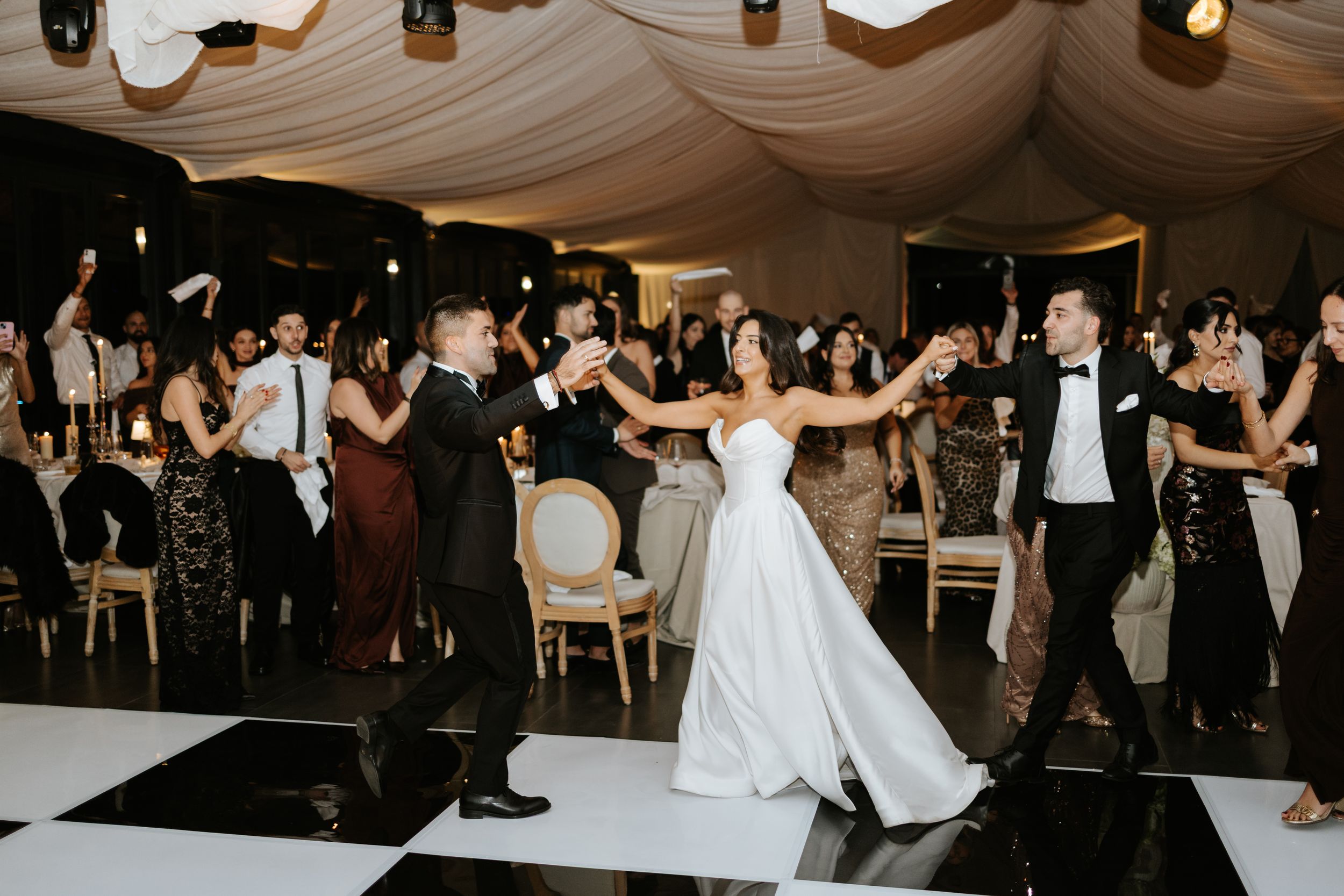 Bride and groom dance joyfully with guests on a black and white checkered dance floor at elegant wedding reception.