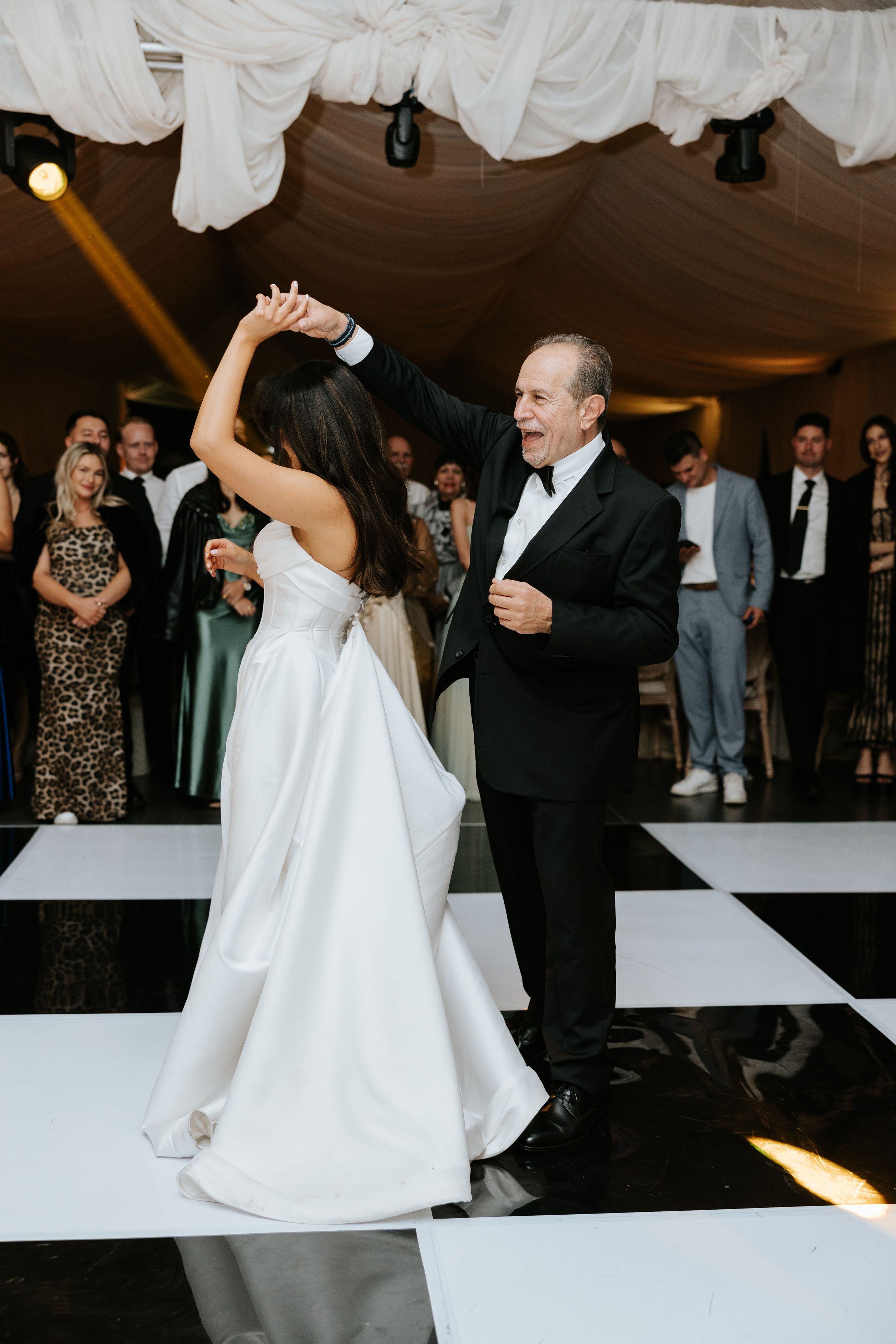 Bride and father share a joyful first dance on black and white checkered floor at elegant wedding reception.