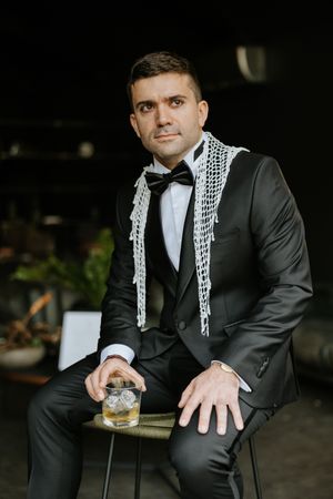 Dapper groom in black tuxedo with bow tie sits holding whiskey glass adorned with white floral garland.