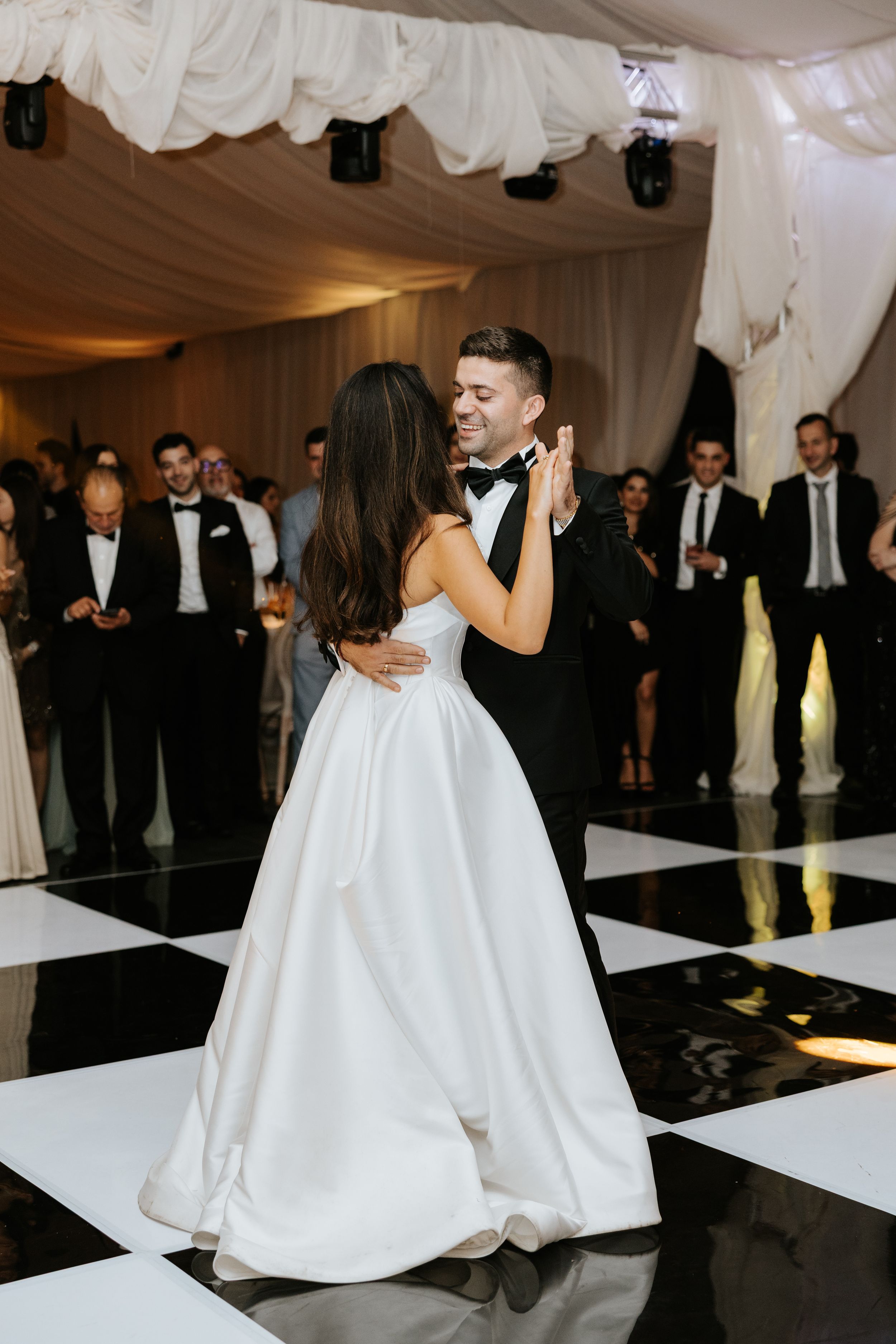 Bride and groom share first dance on black and white checkered floor in elegant draped wedding reception venue.