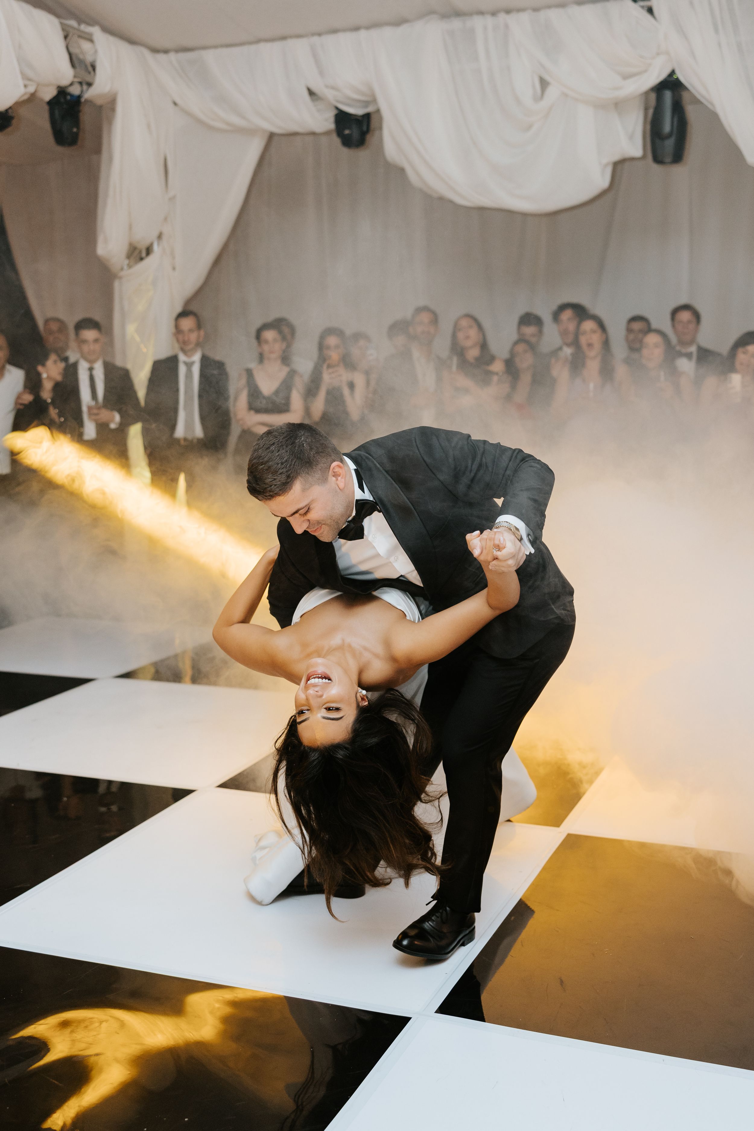 Groom dramatically dips bride on checkered dance floor surrounded by dry ice fog at wedding reception.