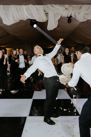 Groom dancing energetically on black and white checkered dance floor at wedding reception with guests watching.