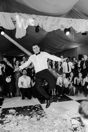 Black and white photo of groom jumping on dance floor surrounded by broken plates at wedding celebration.