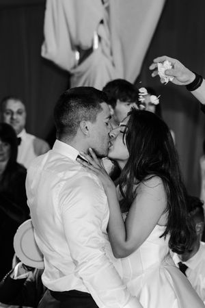 Black and white photo of newlyweds sharing romantic first kiss at wedding reception with guests nearby.