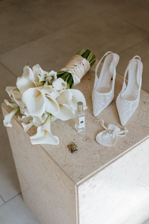 White calla lily bridal bouquet with lace heels, perfume bottle, and accessories arranged on marble surface.