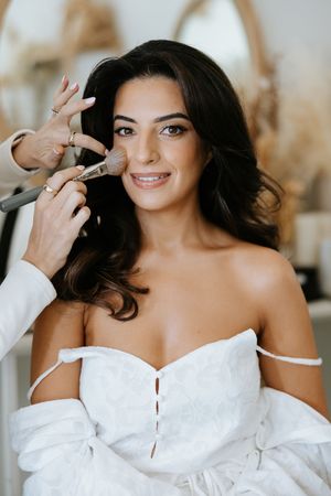 Beautiful dark-haired bride smiling while makeup artist applies blush with brush during wedding preparations.