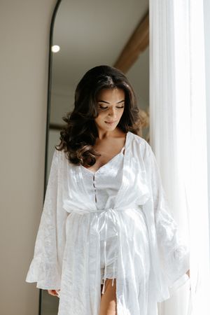 Beautiful dark-haired bride in white lace robe stands by mirror and sheer curtains during wedding morning prep.
