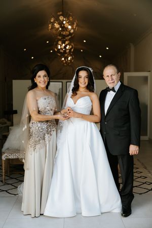 Bride in white ball gown posing with parents under golden chandelier before wedding ceremony.