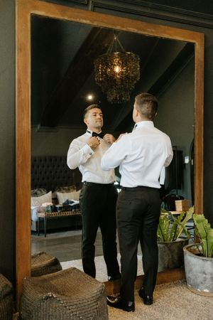 Groom adjusts bow tie while getting ready in front of large wooden mirror at wedding venue.