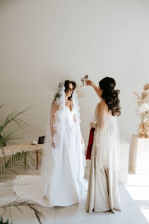 Bridesmaid helps bride with lace veil in bright white room during wedding day preparations.