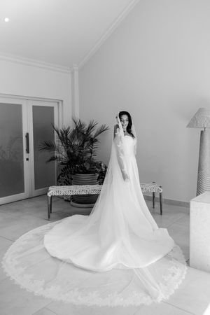 Elegant bride in a long white wedding gown with lace-trimmed cathedral veil posing in a bright room.
