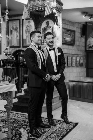Black and white photo of groom and groomsman laughing together inside an Orthodox church before ceremony.