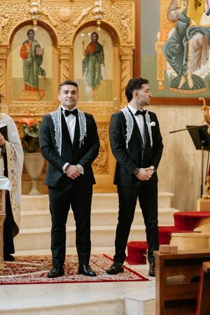 Two groomsmen in black tuxedos with traditional scarves stand at church altar during wedding ceremony.